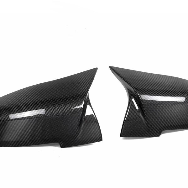 Two carbon fiber mirror covers are positioned side by side, facing each other against a plain white background, reflecting light subtly across their glossy, textured surfaces.