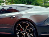 A sleek, metallic gray car, parked with its back wheel in focus, features stylish black alloy rims. Surrounding it are green trees, suggesting a serene, natural environment.