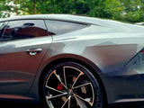 A sleek gray sports car is parked, showcasing its polished body and distinctive alloy wheels. Surrounding trees and greenery slightly blur in the background, emphasizing the car's design.