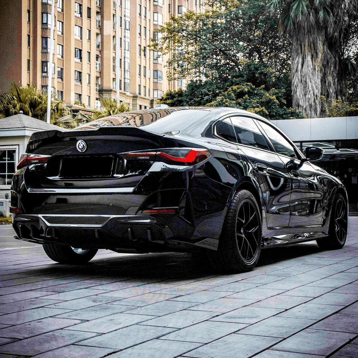 A sleek black car is parked on a tiled sidewalk. Tall buildings and lush trees surround it, creating an urban setting. The detailed design of the car is prominent.
