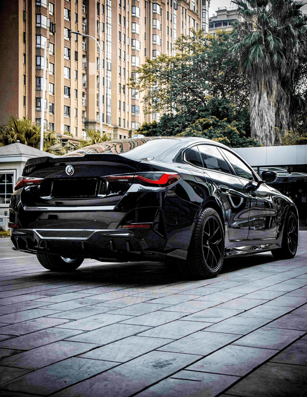 A sleek black car is parked on a tiled sidewalk. Tall buildings and lush trees surround it, creating an urban setting. The detailed design of the car is prominent.