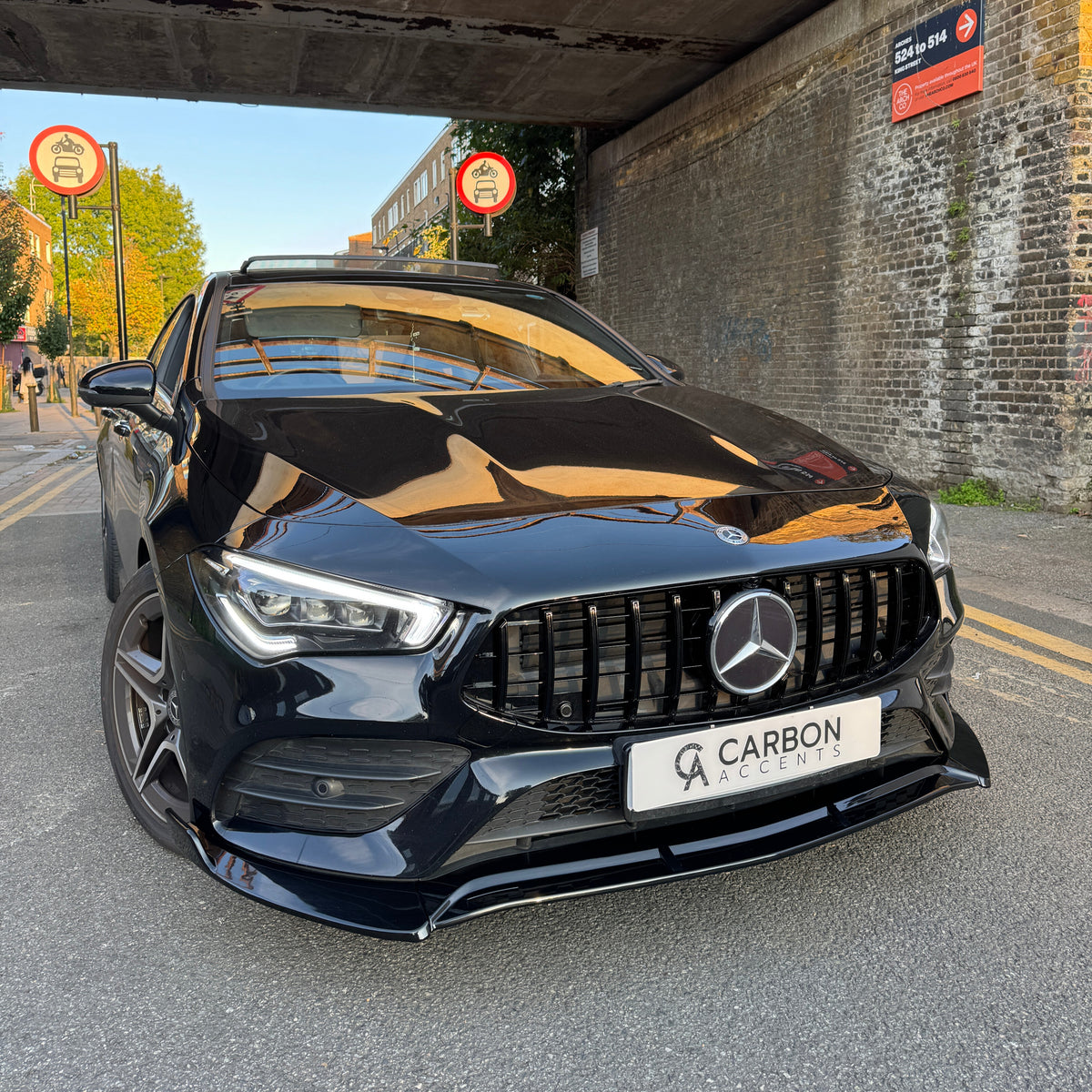 Car: A black Mercedes-Benz, stationary, with a "CARBON ACCENTS" license plate. Context: Parked under a bridge with brick walls, surrounded by "No Cycling" signs and a nearby street with trees and buildings.