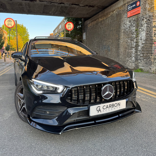 Car parked under a bridge, facing forward. Two "no cycling" signs are visible nearby. License plate reads "CARBON ACCENTS." Brick wall and urban street setting with people in the distance.