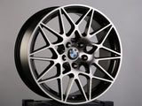 A black and silver BMW alloy wheel with intricate spoke design rests on a dark box. The BMW logo is visible at the center. The background is plain gray. Text on the box has 