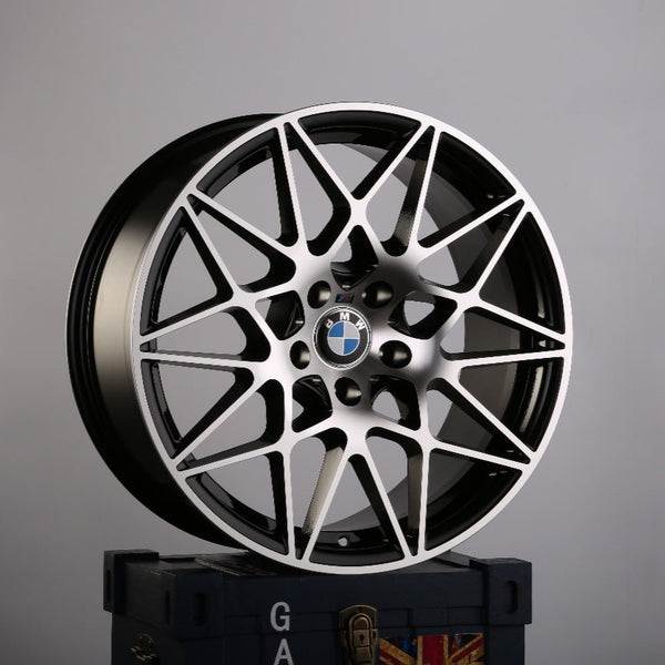 A black and silver BMW alloy wheel with intricate spoke design rests on a dark box. The BMW logo is visible at the center. The background is plain gray. Text on the box has "GA."