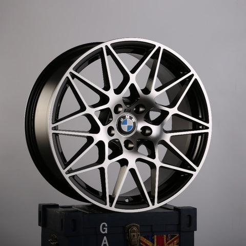 A black and silver BMW alloy wheel with intricate spoke design rests on a dark box. The BMW logo is visible at the center. The background is plain gray. Text on the box has "GA."