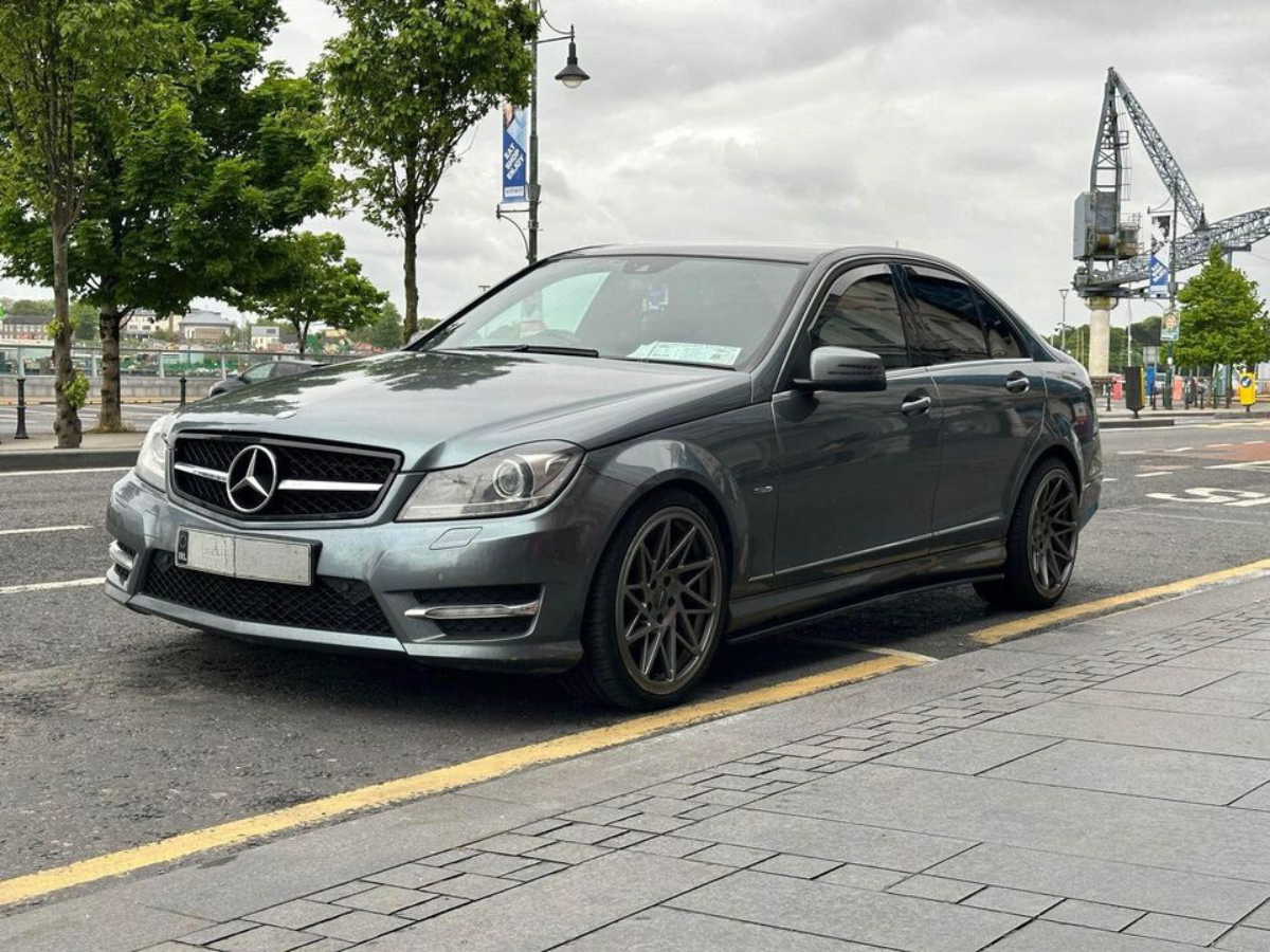 A gray Mercedes sedan is parked on a city street near trees and a crane, under a cloudy sky. There is a license plate visible on the front of the car.