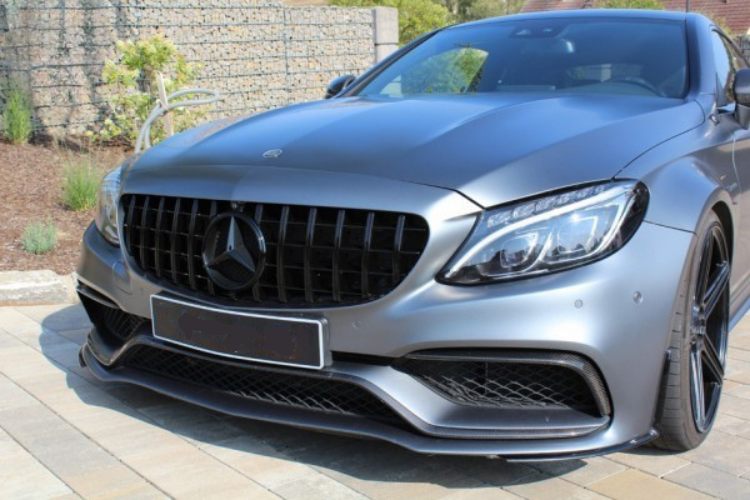 A metallic gray sports car with a prominent grille and sleek headlights is parked on a tiled driveway, surrounded by a neatly landscaped garden and stone wall.