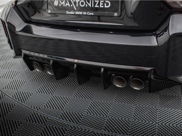 A car's rear bumper displays a custom exhaust with multiple pipes and a diffuser. The scene is set on a geometric-patterned surface. Text: "#MAXTONIZED Dealer BMW M-Cars".