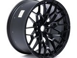 A glossy black alloy wheel with a complex, multi-spoke design is standing upright, highlighting its intricate geometric pattern against a plain white background. A label with text is visible on the rim’s inner surface.