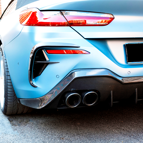 A blue BMW car is parked on a city street. It features dual exhausts and carbon fiber detailing, with other vehicles visible in the background.