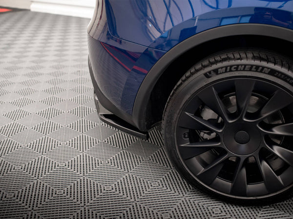 A car's rear bumper and black tire with "MICHELIN 255/40R20" text are parked on a patterned gray surface, reflecting a building's geometric outline.