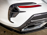 A car's rear bumper features carbon fiber detailing, red tail light accents, and an exhaust outlet, set against a clean, white body in a garage environment.