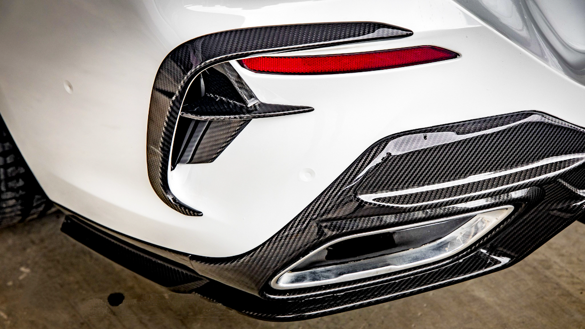 A car's rear bumper features carbon fiber detailing, red tail light accents, and an exhaust outlet, set against a clean, white body in a garage environment.
