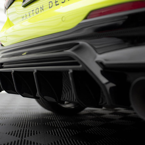 Car rear bumper with a black aerodynamic diffuser, showing multiple fins, in a garage setting with a textured floor. Text reads "MAXTON DESIGN" on the yellow car body.