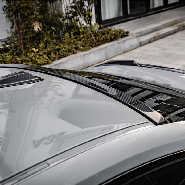 A car with a sleek carbon fiber roof detail reflecting nearby buildings is parked beside a landscaped sidewalk and modern exterior.