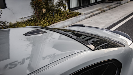 A car with a sleek carbon fiber roof detail reflecting nearby buildings is parked beside a landscaped sidewalk and modern exterior.