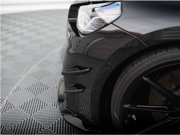 A black car's front corner, including the headlight and tire, is shown on a checkered-patterned floor, emphasizing its sleek design and glossy finish.