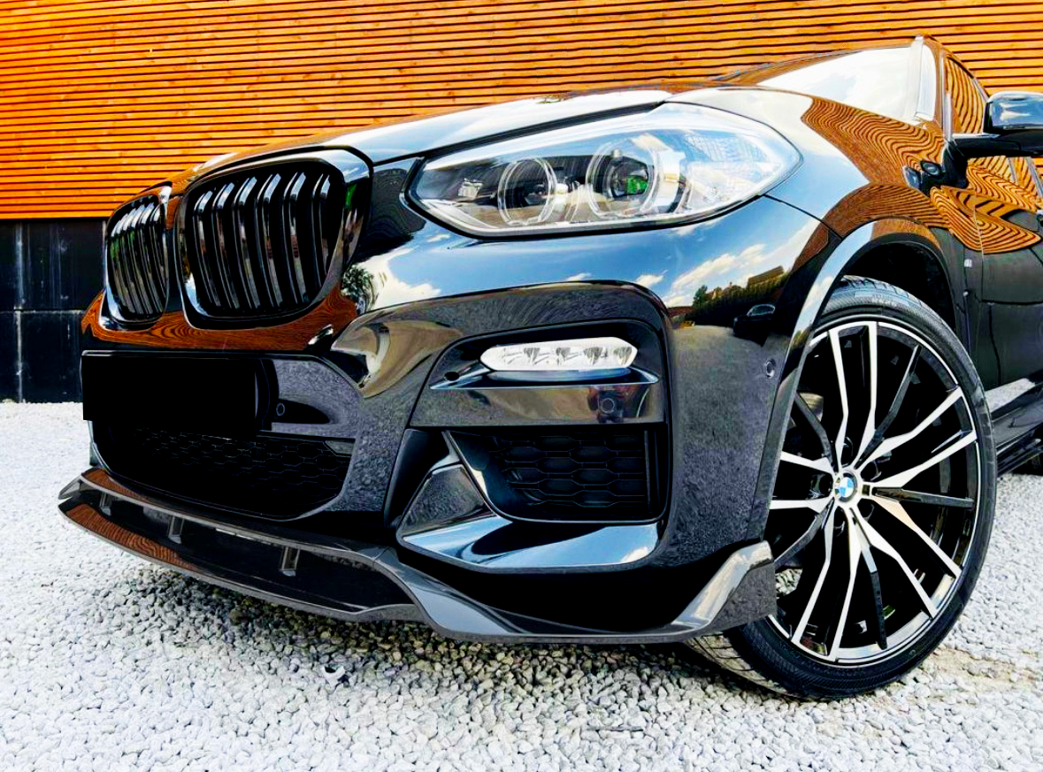 A black car with sleek headlights and multi-spoke alloy wheels is parked on white gravel in front of a wooden-paneled wall, reflecting surroundings on its glossy surface.