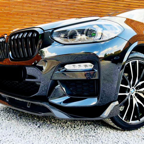 A black car with sleek headlights and multi-spoke alloy wheels is parked on white gravel in front of a wooden-paneled wall, reflecting surroundings on its glossy surface.