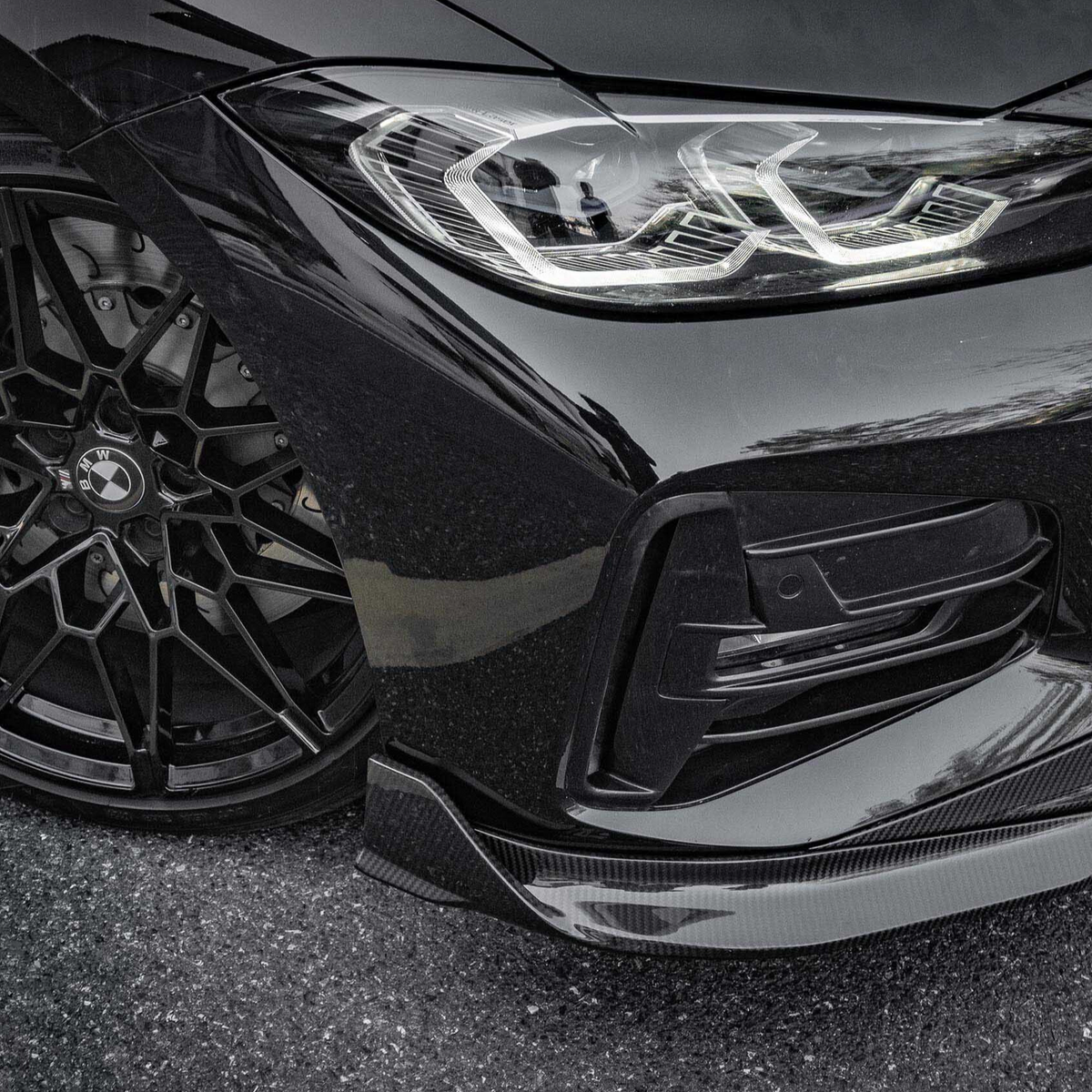 Black car with intricate alloy wheel design and sleek headlight, parked on an asphalt surface. Reflections highlight the car's glossy finish. BMW badge visible on the wheel hub.