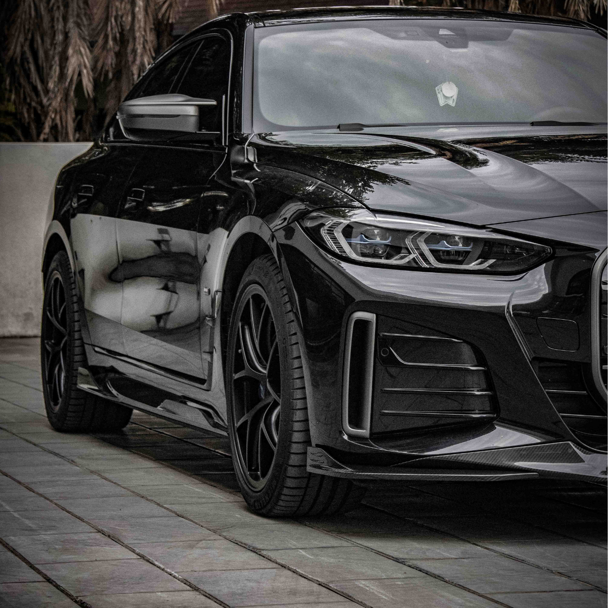 A sleek black car, parked on a tiled surface, reflecting surrounding trees and buildings under a cloudy sky, with palm leaves visible in the background.