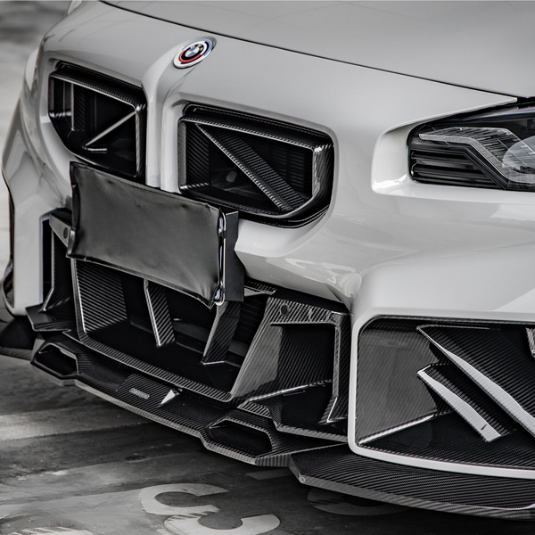 A white car with a sporty front bumper featuring intricate carbon fiber accents, parked in a concrete garage. A covered license plate is visible in the center.