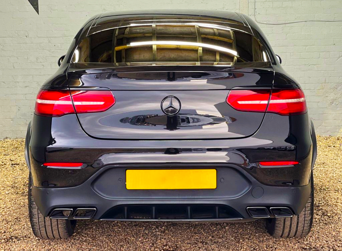A black car, featuring glowing red taillights and a rear license plate, is parked on gravel against a light brick wall. It displays a Mercedes-Benz logo on the trunk.
