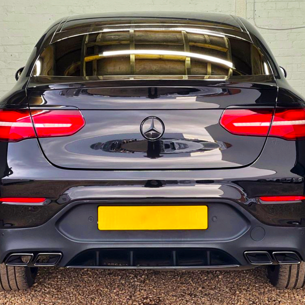 A black car, featuring glowing red taillights and a rear license plate, is parked on gravel against a light brick wall. It displays a Mercedes-Benz logo on the trunk.