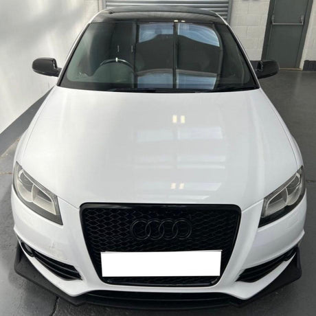 A white Audi car is parked in a garage, viewed from the front, displaying its black grille with the Audi logo prominently in the center. The environment is industrial.