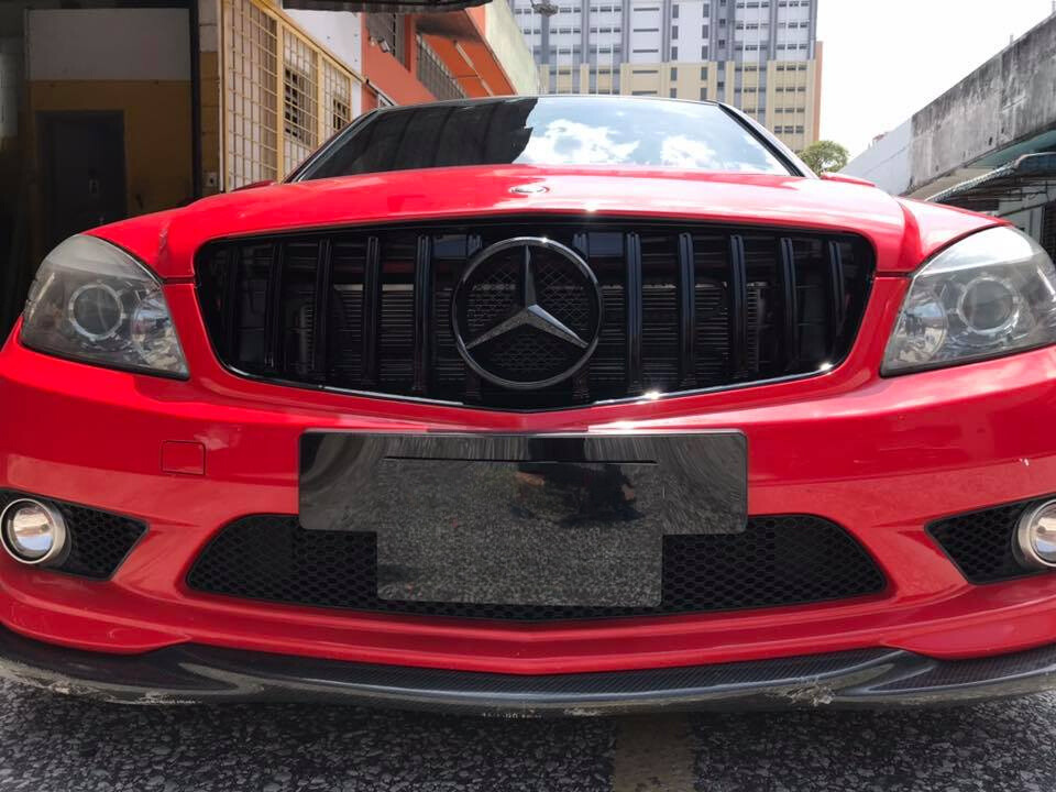A red Mercedes-Benz car is parked in an urban environment, displaying a black grille with a prominent Mercedes emblem and surrounded by buildings.