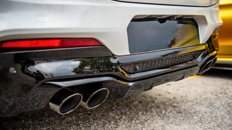 The car's rear features a sleek black bumper with dual exhaust pipes, framed by a white body and red tail light, parked on a gray asphalt surface beside another vehicle.