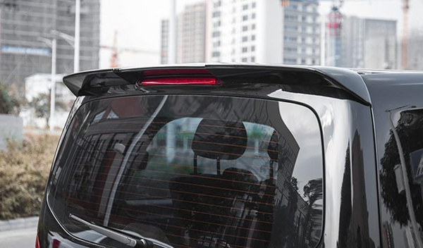 A black vehicle's rear window reflects surrounding trees and buildings, with a red brake light on top. In the background, urban buildings and construction cranes are visible.