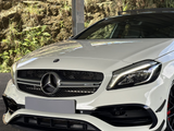 White Mercedes-Benz car sits parked under a metal structure; its front grille features the Mercedes logo and “AMG” badge; lush greenery and rocky terrain are visible in the background.