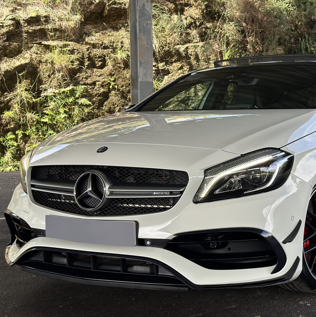 White Mercedes-Benz car sits parked under a metal structure; its front grille features the Mercedes logo and “AMG” badge; lush greenery and rocky terrain are visible in the background.