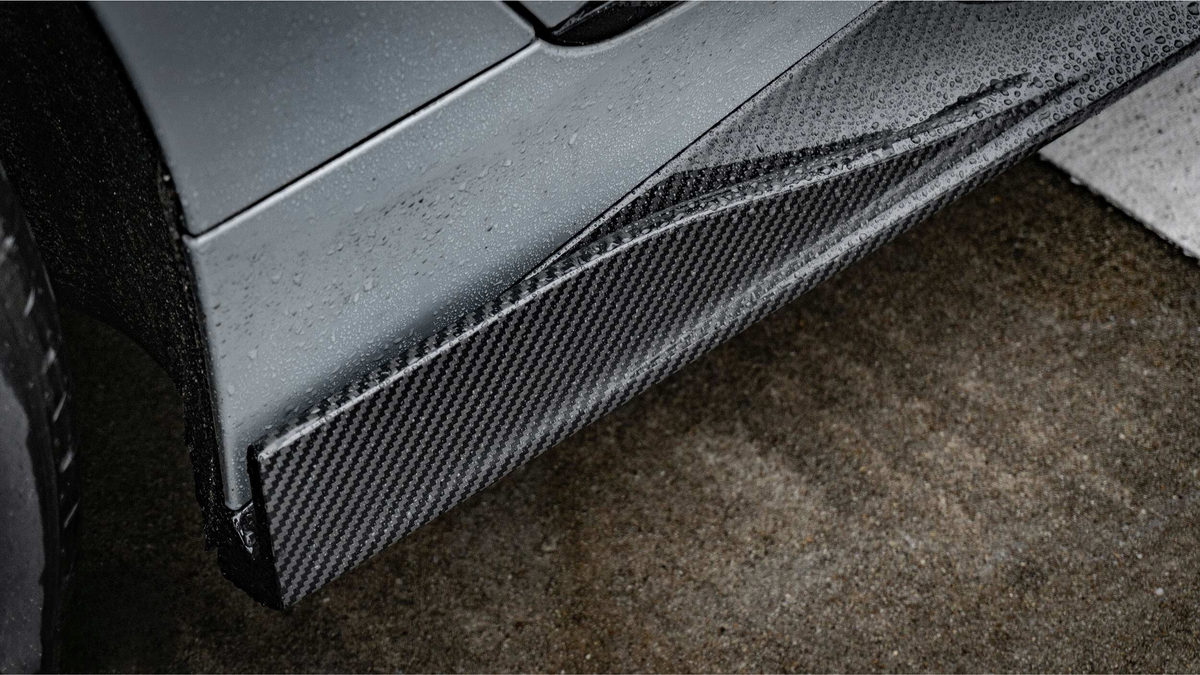 A sleek car's carbon fiber side skirt, covered in raindrops, sits adjacent to a wet asphalt surface, highlighting the vehicle's modern design and performance-oriented components.