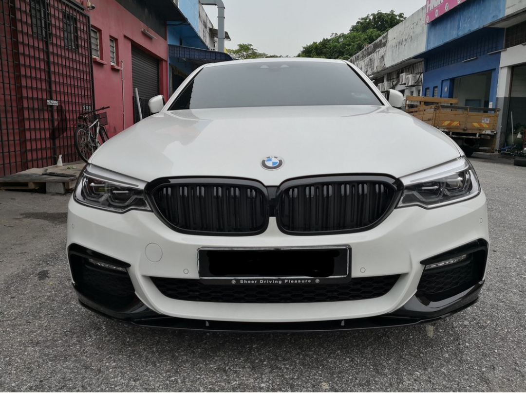 A white BMW car is parked on a gravel surface between industrial buildings. Text on the license plate area reads "Sheer Driving Pleasure." A bicycle and wooden pallets are nearby.