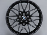 A black BMW alloy wheel with intricate spoke design is displayed vertically on a circular platform. The center features the BMW logo. The background is plain and light gray.