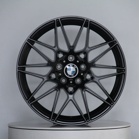A black BMW alloy wheel with intricate spoke design is displayed vertically on a circular platform. The center features the BMW logo. The background is plain and light gray.