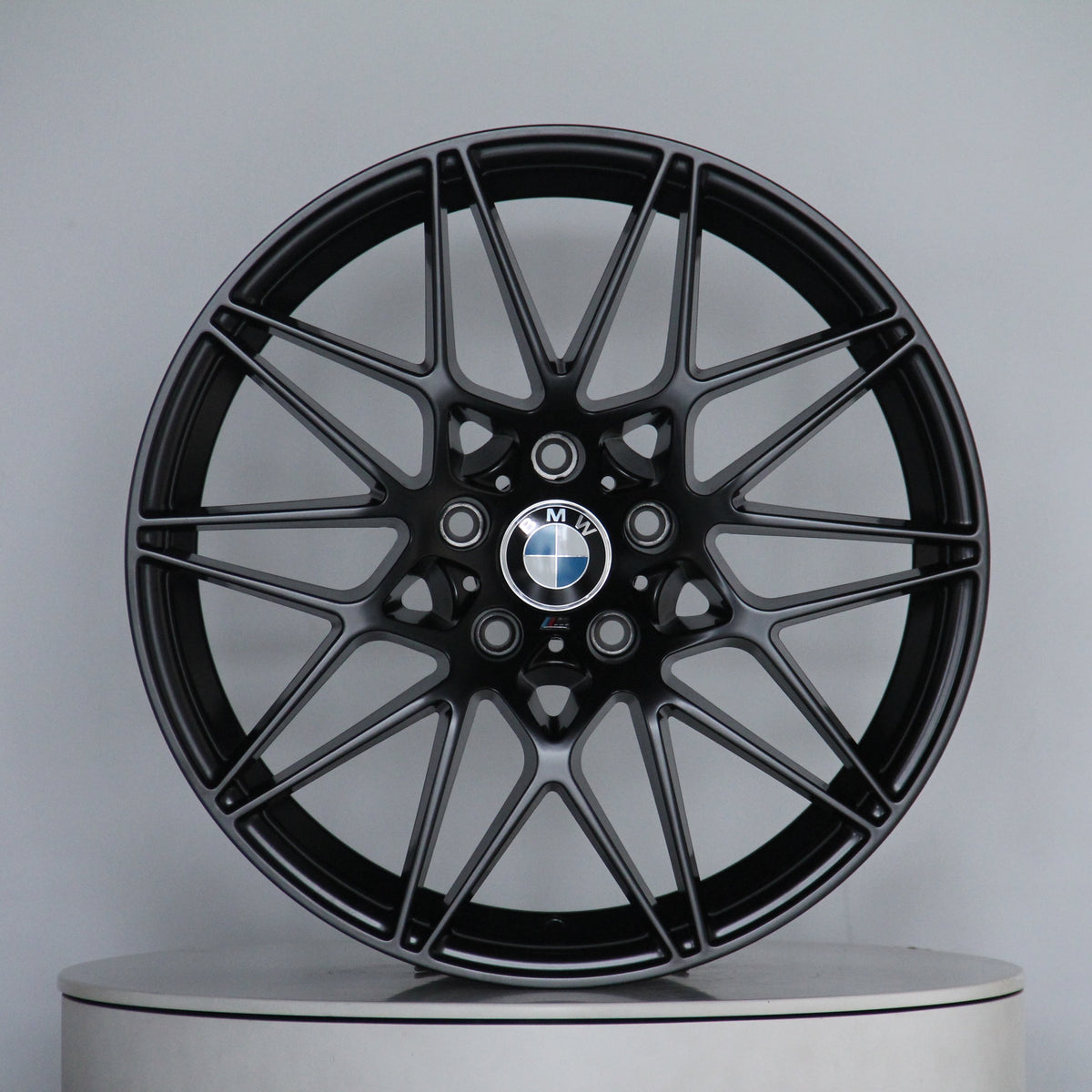 A black alloy wheel with multi-spoke design, featuring a central BMW logo, rests on a round display platform against a plain gray background.