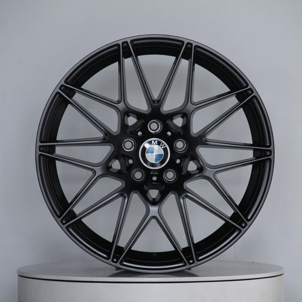 A black alloy wheel with intricate spoke design displays a BMW logo at the center, resting on a round platform against a plain gray background.