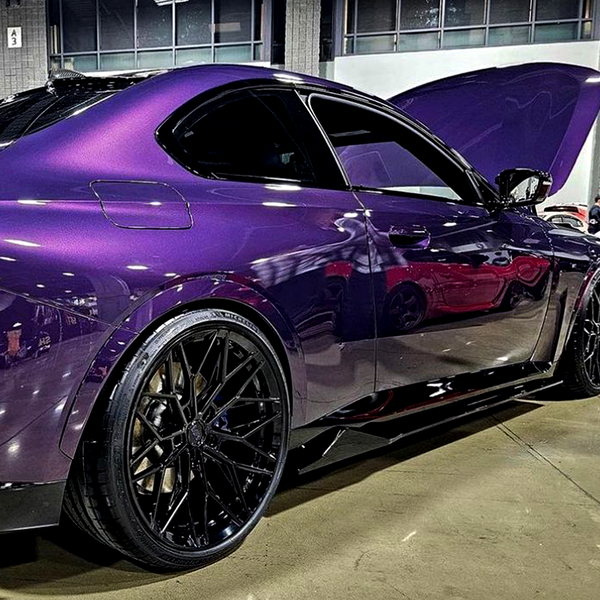A sleek, purple sports car with a raised hood is displayed indoors on a polished concrete floor, featuring glossy black wheels.