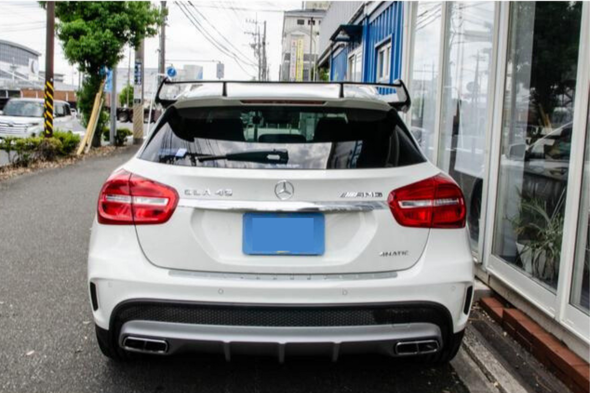 A white Mercedes-Benz GLA 45 AMG is parked on a street, with a distinctive spoiler. Nearby are trees, parked cars, and a blue building. Surrounding cables are visible in the sky.