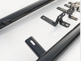 Black metal brackets are lying flat with holes for mounting, alongside elongated cylindrical bars, on a smooth white surface, suggesting parts for assembly or installation.