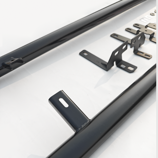 Black metal brackets are lying flat with holes for mounting, alongside elongated cylindrical bars, on a smooth white surface, suggesting parts for assembly or installation.