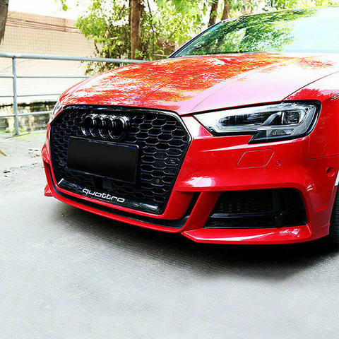 A red car with a prominent honeycomb grille displaying the word "quattro" is parked. The scene features greenery in the background under natural daylight.