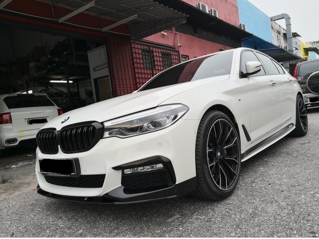A white BMW sedan is parked on a gray asphalt surface outside a garage, surrounded by several other cars and colorful building walls.