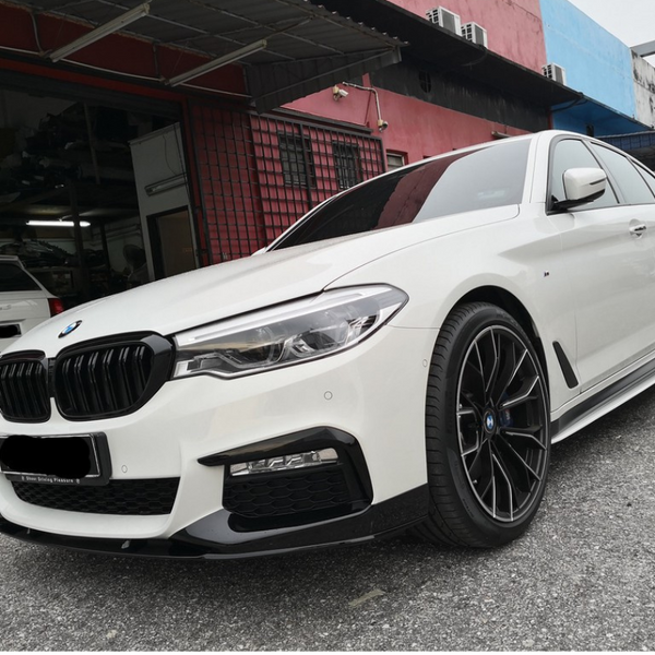 A white BMW sedan is parked on a gray asphalt surface outside a garage, surrounded by several other cars and colorful building walls.