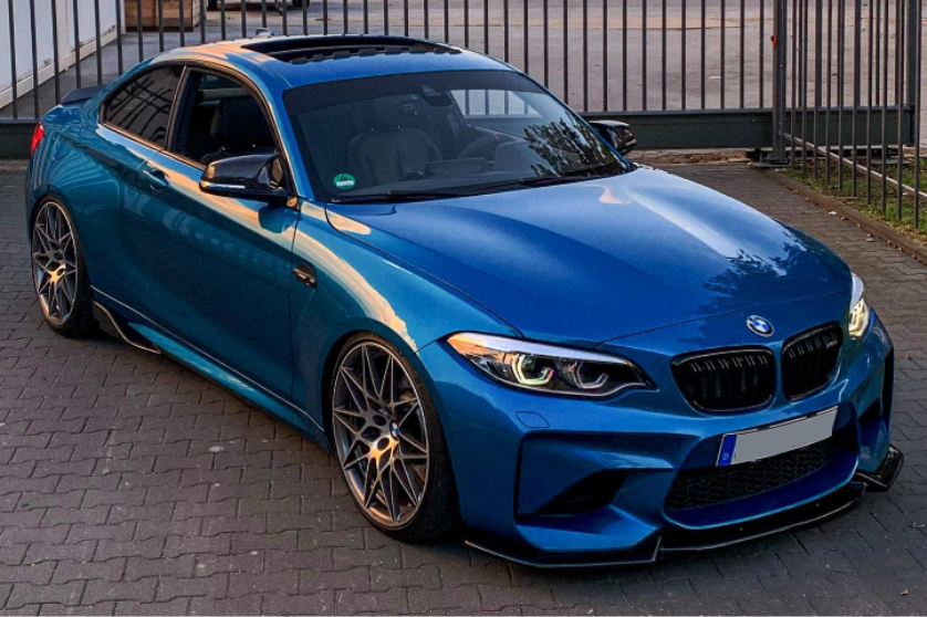 A blue car is parked on a brick driveway, featuring sleek design and large alloy wheels, with a metal fence and building in the background. The license plate is obscured.