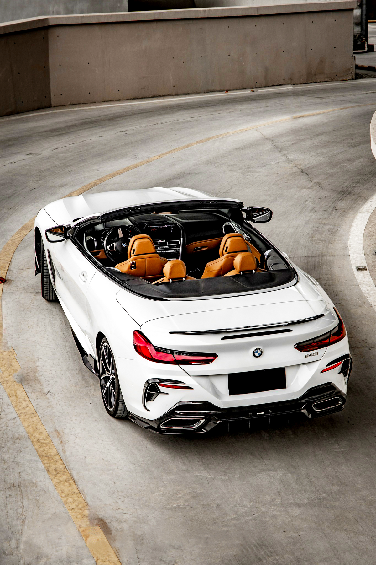 Convertible car is parked on a curving concrete road. The vehicle is white with a black soft top, showcasing tan leather seats. The surrounding area appears to be an industrial parking garage.